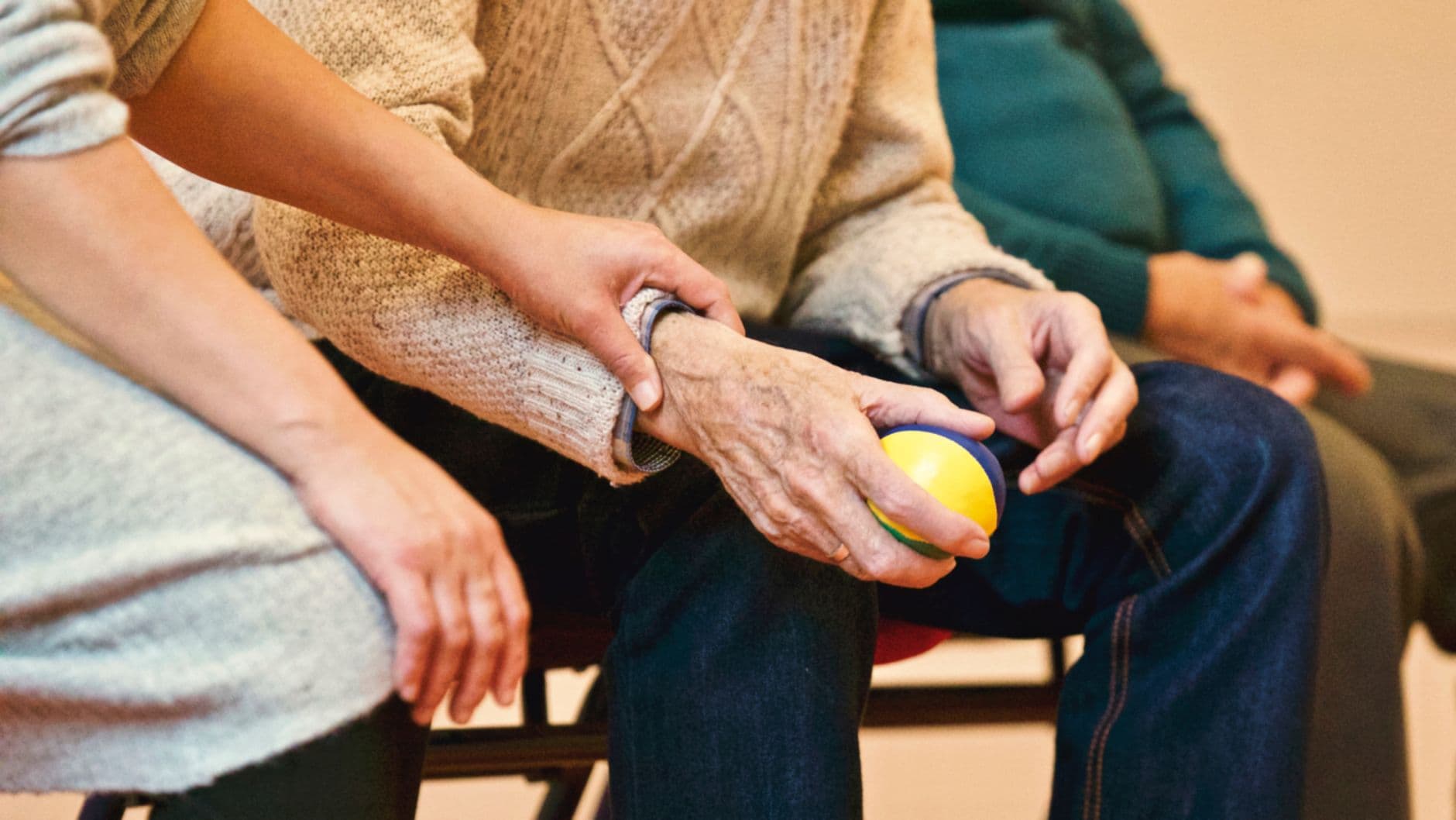Une aidante soutient l'avant-bras d'une personne âgée assise, qui tient une balle de rééducation dans la main.