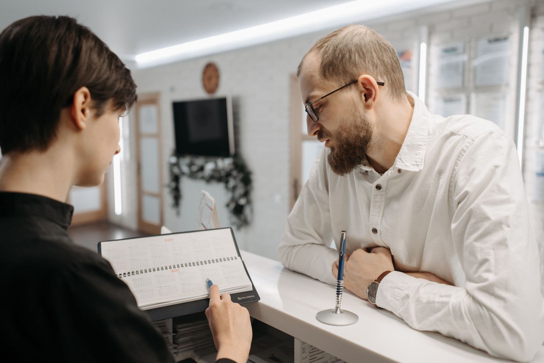 Une secrétaire médicale montre un agenda de rendez-vous à un patient, au comptoir d'accueil d'un cabinet.