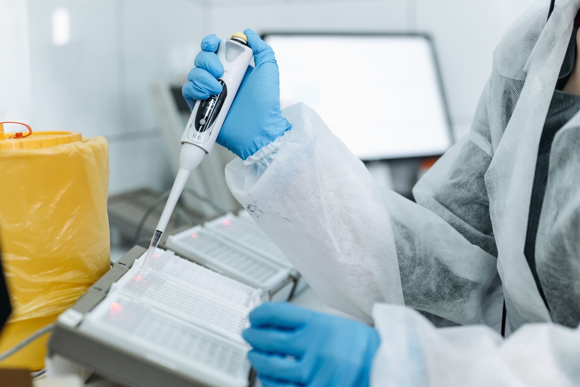 Un technicien de laboratoire en blouse stérile et gants prélève un échantillon à la micropipette sur une plaque d'analyse.