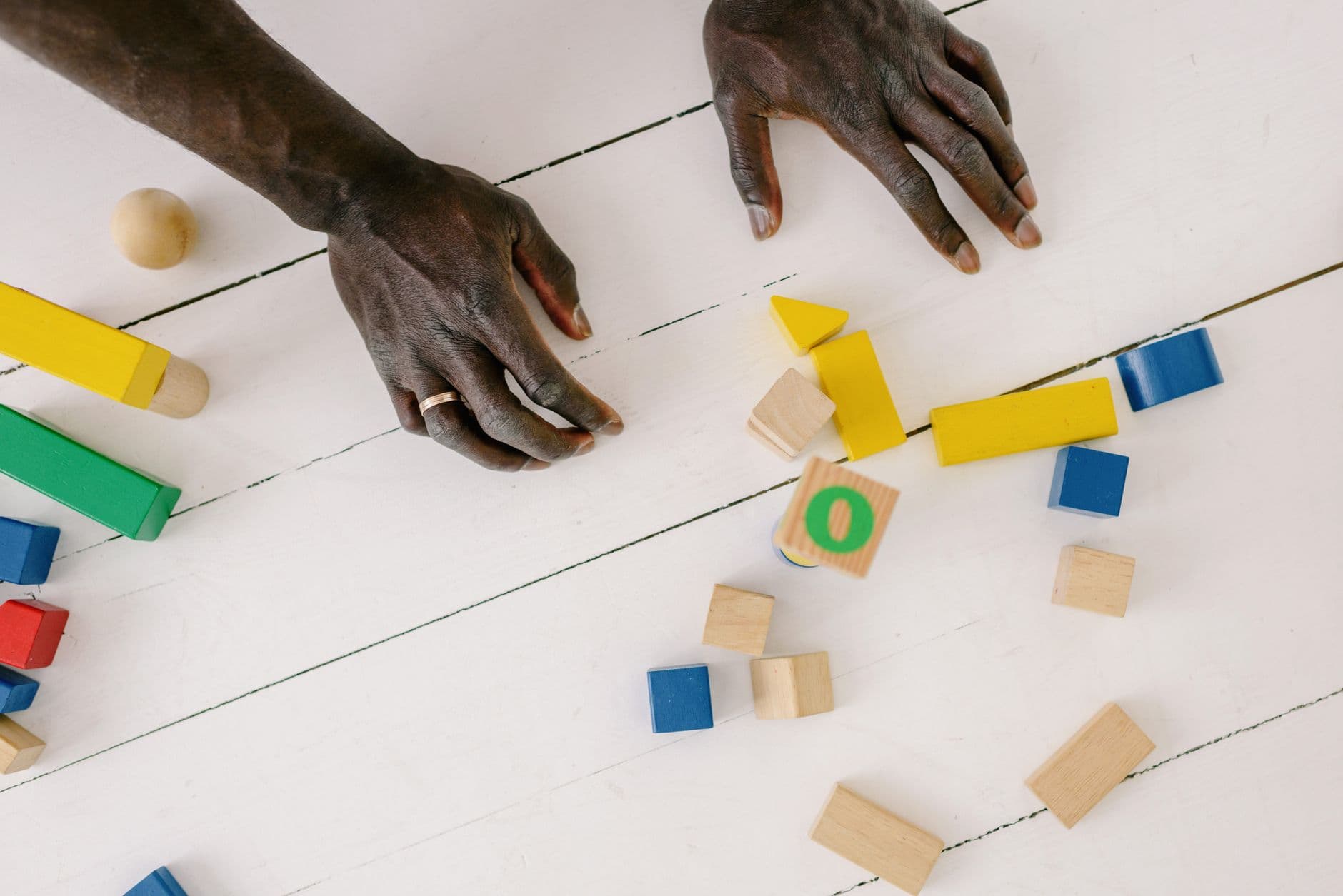 Des mains d'adulte disposent des cubes de bois colorés sur un plan de jeu, vues de dessus.
